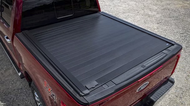 Red truck with a closed black bed cover parked on gravel, featuring a tailgate emblem.