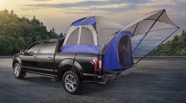 Black pickup truck with a blue tent set up in its bed, set against a forest and sunset sky.
