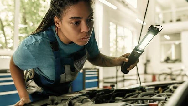 Mechanic inspecting car engine with work light in a garage, wearing a blue shirt and overalls.