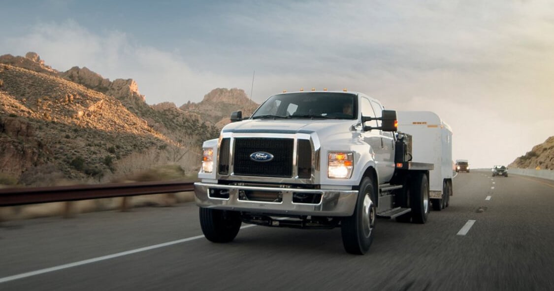 White truck towing a trailer on a highway by scenic mountains under a cloudy sky.