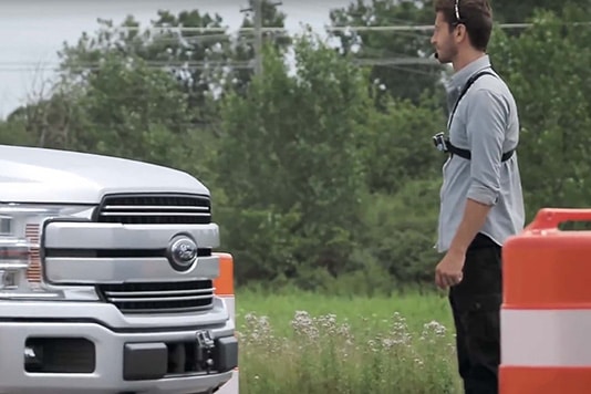 A person stands beside a silver pickup truck on a road lined with orange barriers. Trees are in the background.