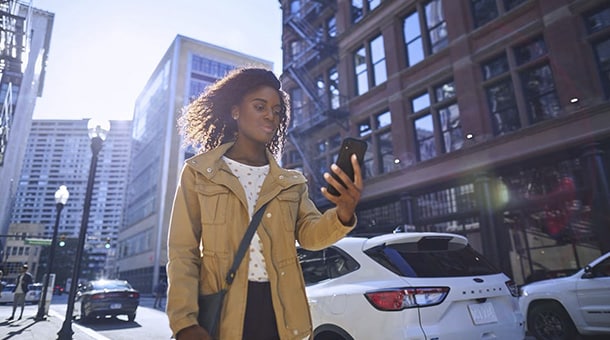 Woman walking in city street, looking at smartphone, urban buildings and cars in background.