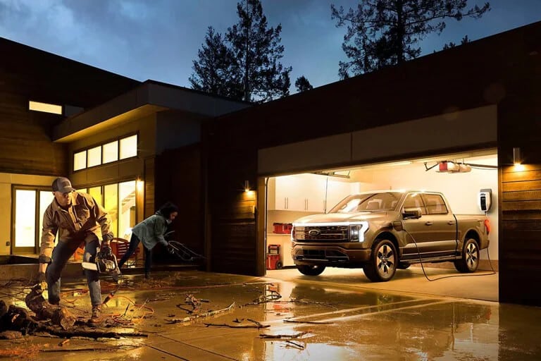 Man using a chainsaw outside a modern house, while a pickup truck charges in the garage.