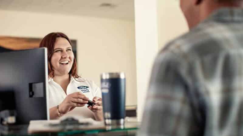 A smiling receptionist assists a customer at a service desk, holding a car key and conversing.