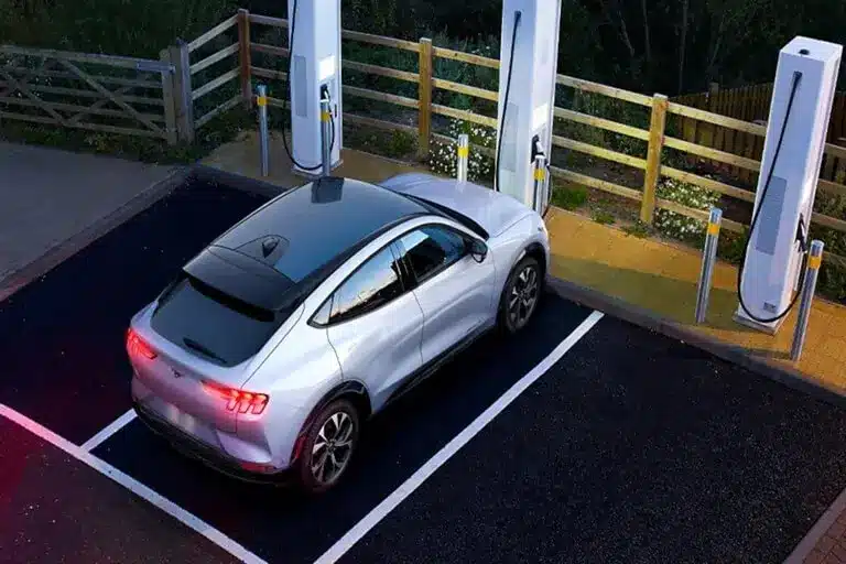 Electric car charging at a station, parked between two charging units with a wooden fence nearby.