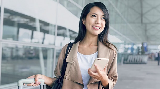 Woman at airport with suitcase and phone, smiling and looking forward.