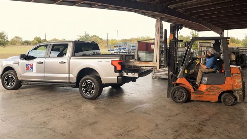 A forklift loads materials onto a silver pickup truck in a warehouse setting.