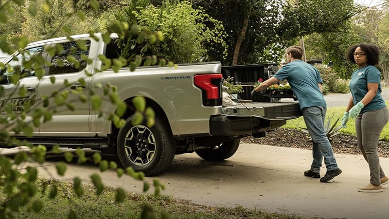 Two landscapers unloading plants from a truck on a driveway, surrounded by greenery.
