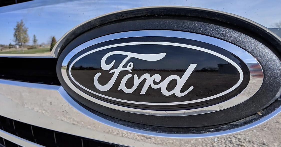 Close-up of a Ford logo on a car grill, with outdoor scenery reflected in the chrome.