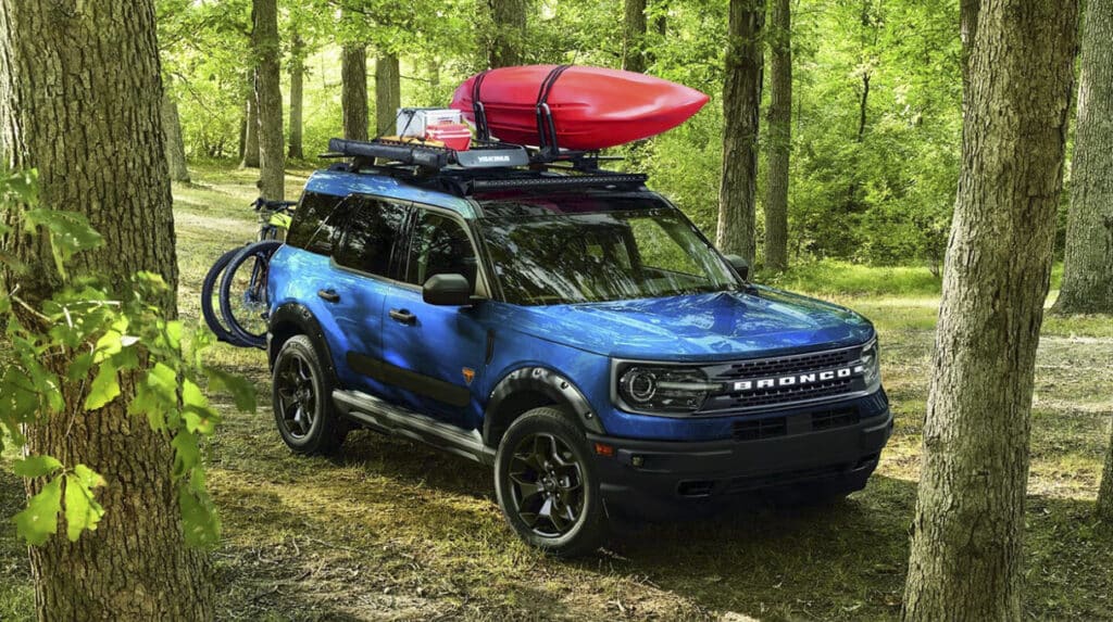 A blue SUV with a red kayak on the roof and a bike attached, parked in a lush green forest.