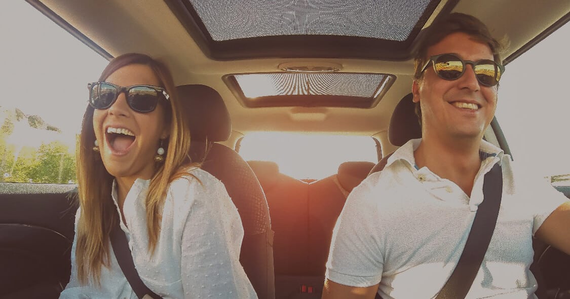 Couple smiling in a car with sunglasses on, enjoying a sunny day drive.