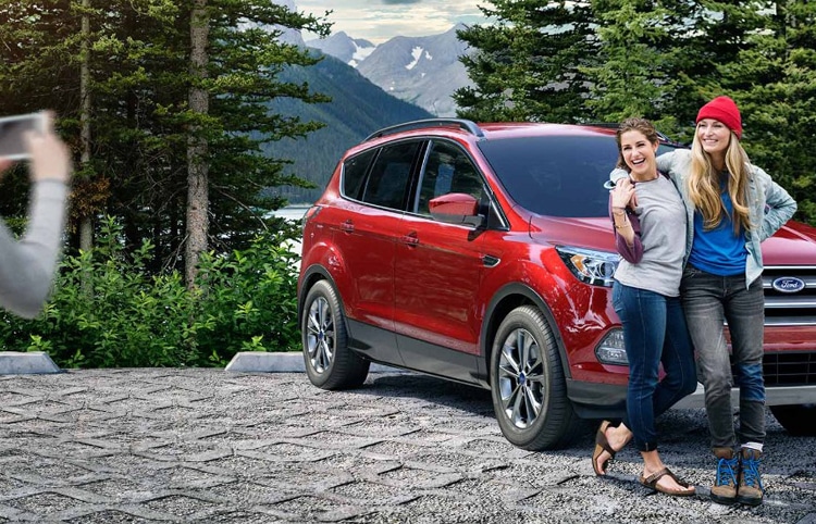 Two women posing with a red Ford SUV in a scenic mountain setting, capturing a photo.