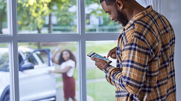 Man in plaid shirt using smartphone indoors, with woman entering a white car outside.