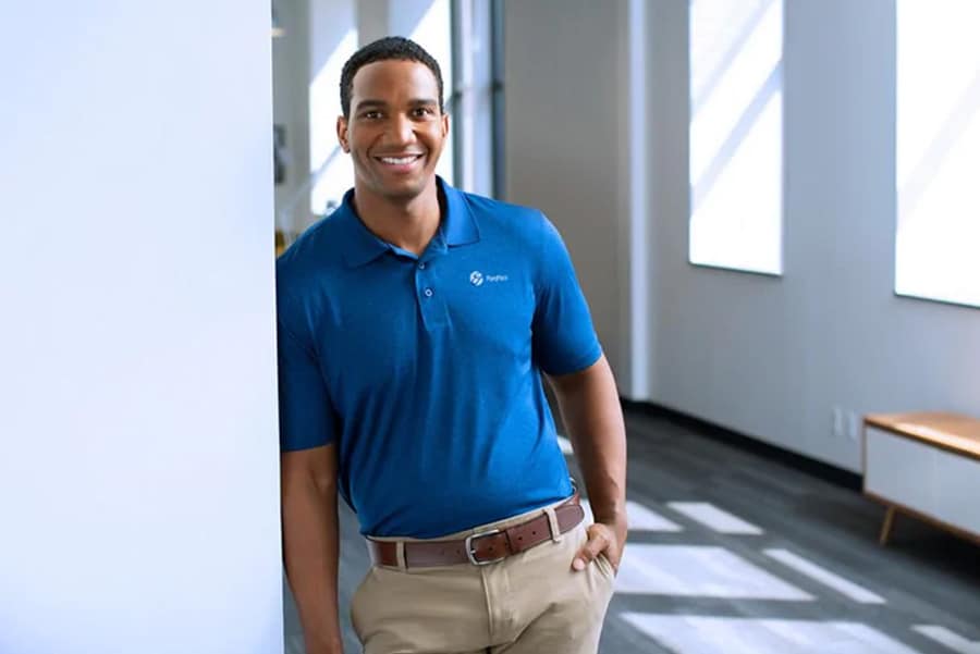 Homme souriant en polo bleu appuyé contre un mur dans un bureau moderne éclairé.