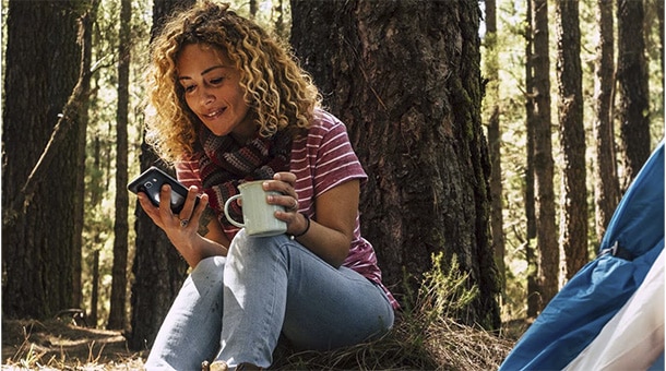 Femme souriante, vêtue d'un t-shirt rayé, utilise un smartphone en tenant une tasse, assise près d'une tente dans la forêt.