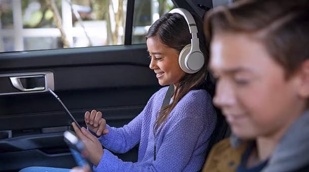 Une fille portant des écouteurs utilise une tablette dans une voiture, souriante. Un garçon est à côté avec un smartphone. Idéal pour le divertissement en voyage.