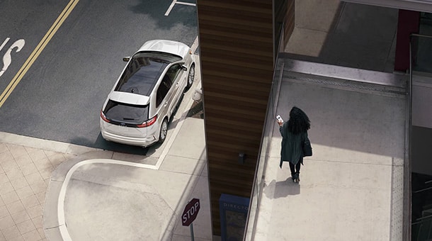 Une voiture blanche est garée près d'un trottoir dans une rue de ville. Une personne marche en regardant son téléphone.