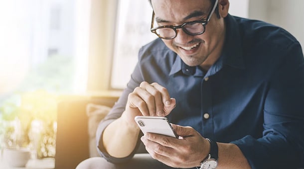 Un homme souriant utilise un smartphone, assis dans un espace lumineux avec des lunettes et une chemise bleue.