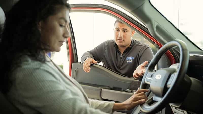 Un homme discute avec une femme à l'intérieur d'une voiture rouge, expliquant probablement une fonctionnalité automobile.
