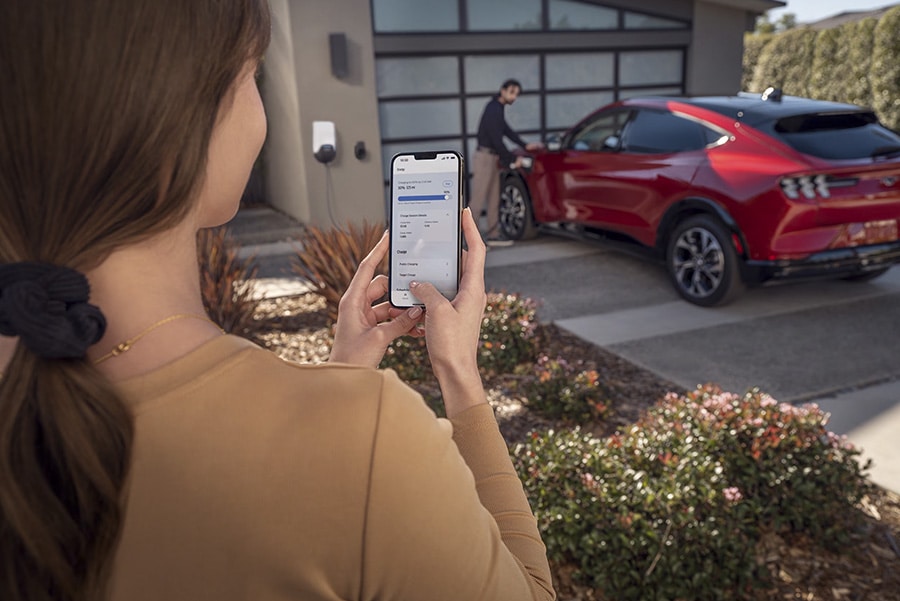 Femme utilisant un smartphone pour gérer la charge d'une voiture électrique rouge garée devant un garage moderne.