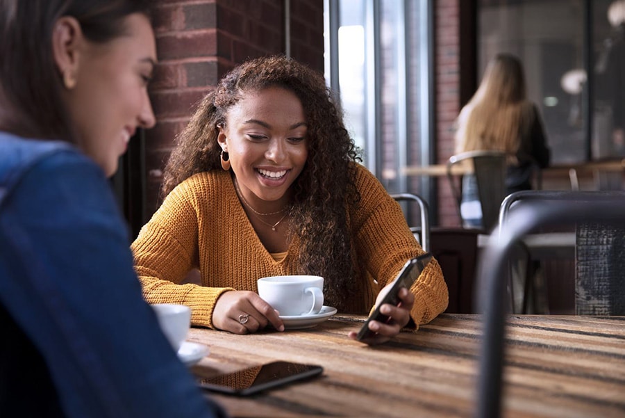 Deux femmes souriantes prennent un café dans un café, échangeant des messages sur un smartphone. Ambiance conviviale.