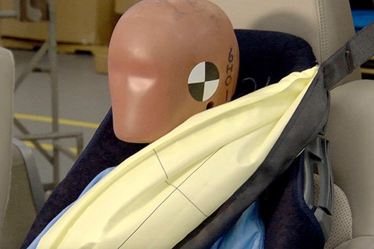 Mannequin de crash-test avec ceinture et airbag déployé dans un siège auto, simulant un impact.