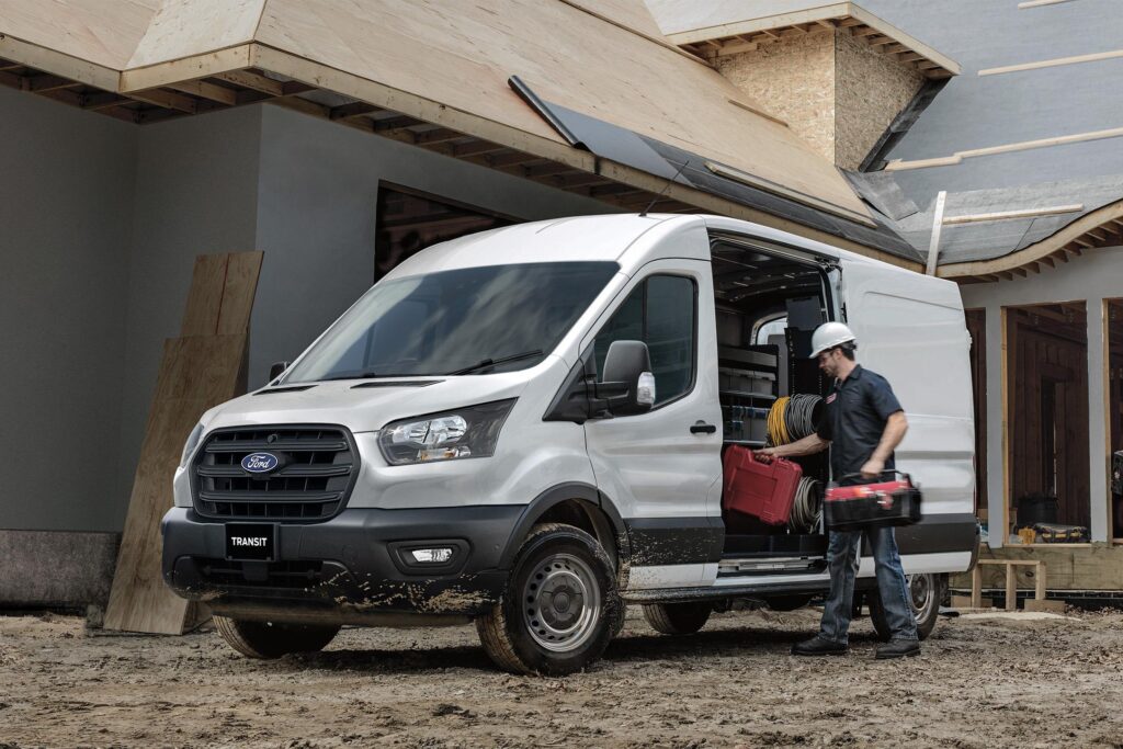 Un fourgon blanc Ford Transit est garé sur un chantier. Un homme en casque blanc charge du matériel.