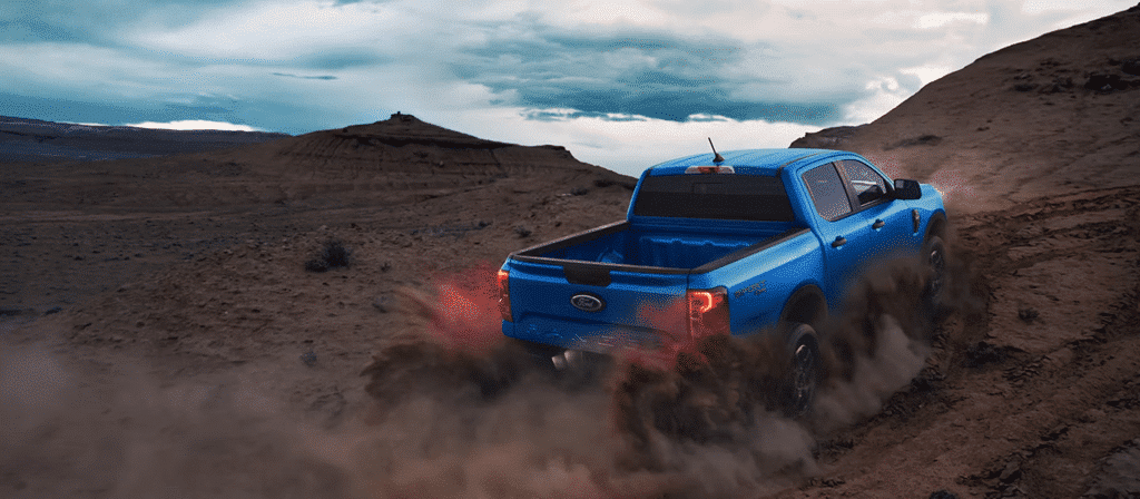 Pick-up bleu roulant sur un terrain accidenté désertique, soulevant une nuée de poussière sous un ciel nuageux.