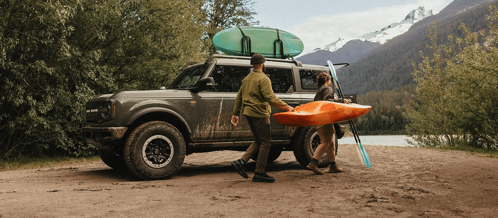 Deux personnes transportent un kayak orange près d'un SUV avec des pagaies et un paddle, en bord de lac montagnard.